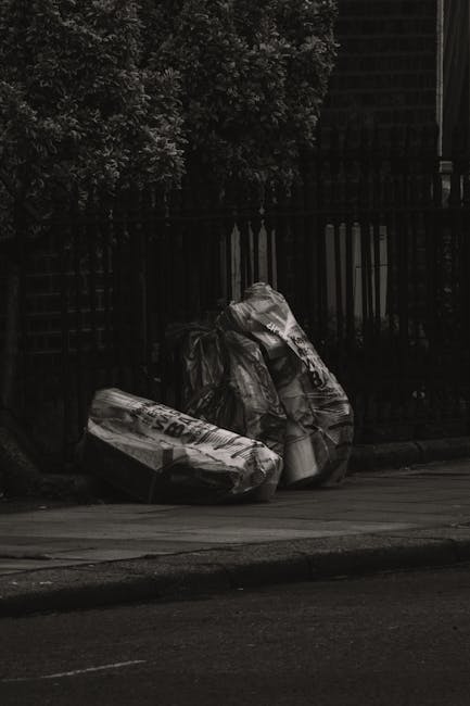 A person in casual clothing is crouching on a paved surface, holding a small wooden stick with a partially broken white plastic container attached to it. The container appears to be a disposable cup o
