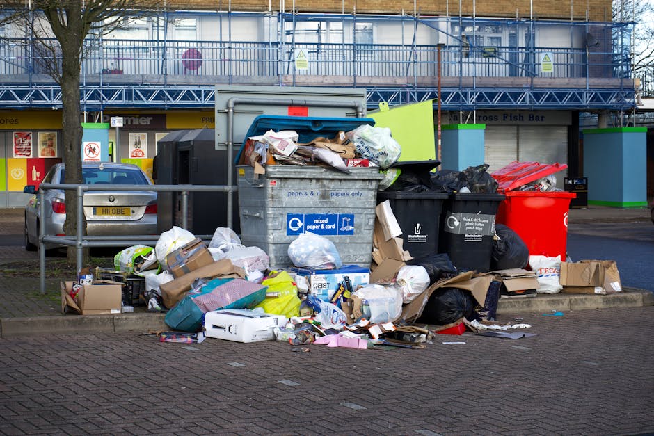 A cluster of overflowing waste bins and overflowing rubbish spilling onto the paved ground of a parking area outside a commercial building. The scene includes a large grey mixed waste bin with a parti