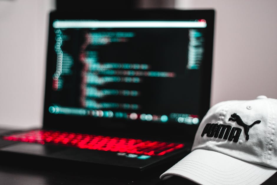 A man with dark hair, glasses, and a beard is sitting at a wooden desk working on a computer. He is using a black keyboard with his hands positioned over it and is looking at two large computer monito