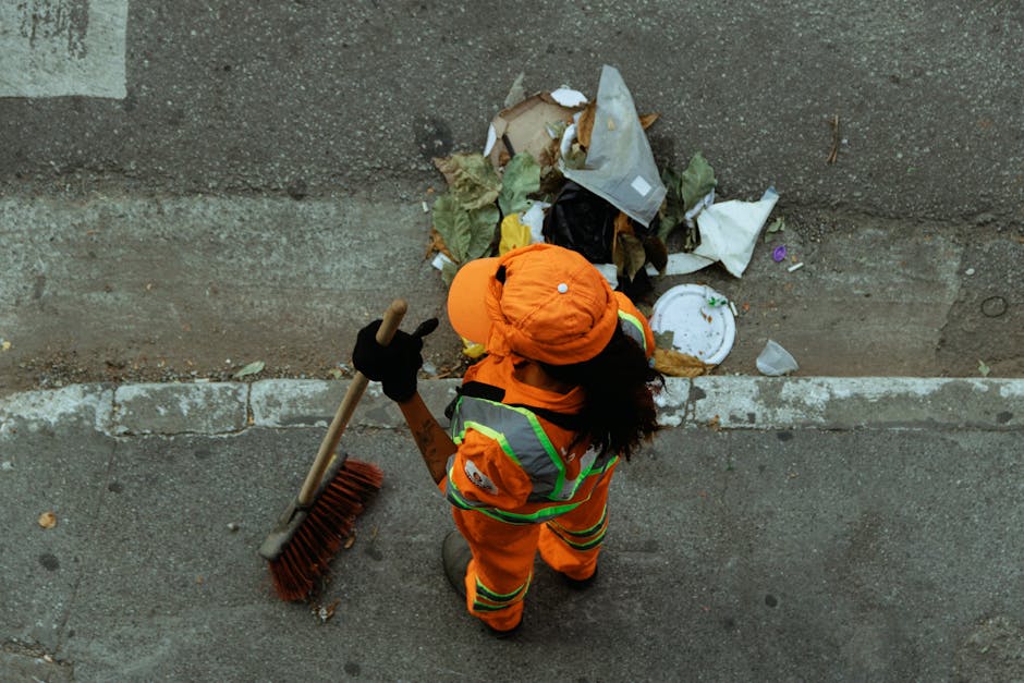 A city street scene featuring a red waste collection truck with its back open, showing mechanical and hydraulic components. A worker wearing a yellow high-visibility vest and black trousers is positio